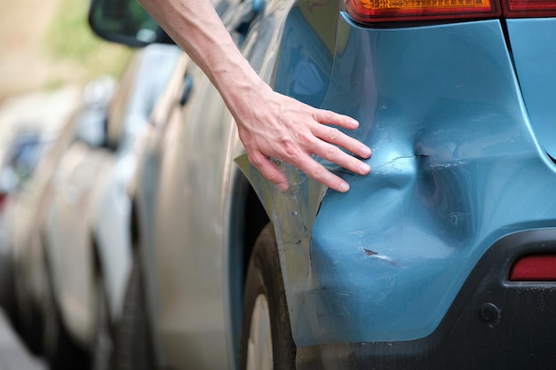 Driver hand examining dented car with damaged fender parked on city street side. Road safety and vehicle insurance concept.