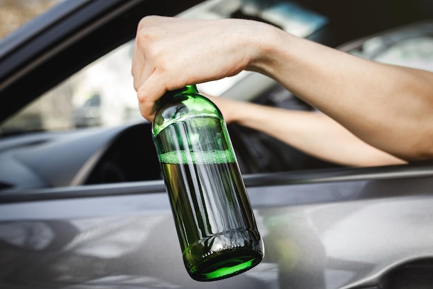 The driver's hand in the car window holds a bottle of alcohol