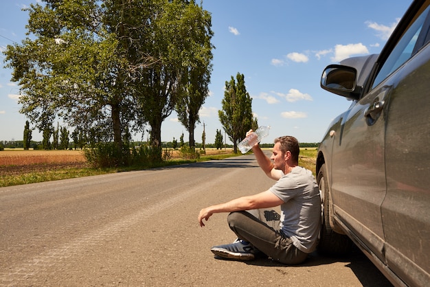 The driver sits on the road near a car with an open hood with a bottle of water on a hot sunny day.