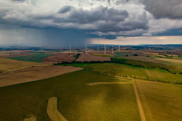 Drone flight at high altitude over a large field with many wind turbines spinning The wind turbines cast a long shadow because the sun is setting In the background at storm sky