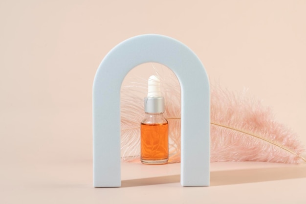 A dropper bottle with an orange nail cuticle oil standing in an arch in front of the pink feather on a beige background