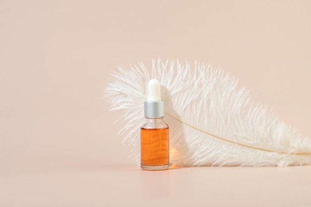 A dropper bottle with an orange nail cuticle oil standing in front of the white feather on a beige background