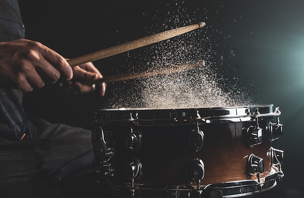 Drummer using drum sticks hitting snare drum with splashing water on black background under studio lighting close up.