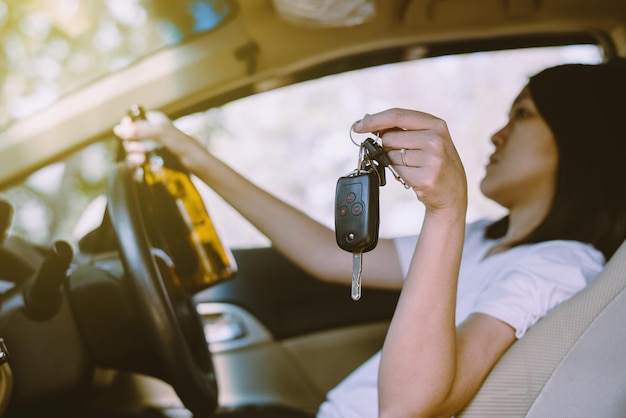 Drunk asian woman holding car key and drinking alcohol while driving car on roadWomen drive her car with a bottle of beerDangerous driving concept