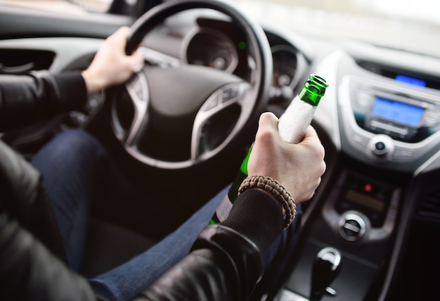 Drunk young man with a beard with a bottle of beer in his hand behind the wheel of a car. violation of law, drunk driving