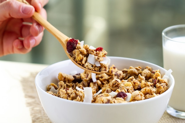 Dry breakfast cereals. Crunchy honey granola with flax seeds, cranberries and coconut in a white bowl and a glass of milk on a table. Healthy and fiber food 