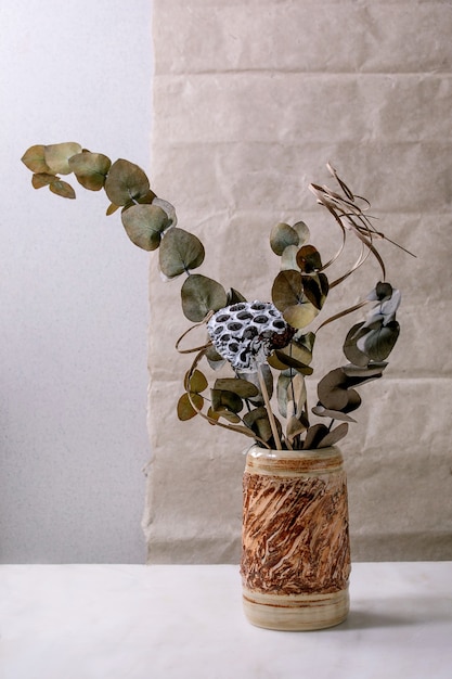 Dry flowers, eucalyptus and twigs branch in brown ceramic vase on white marble table with gray wall behind.