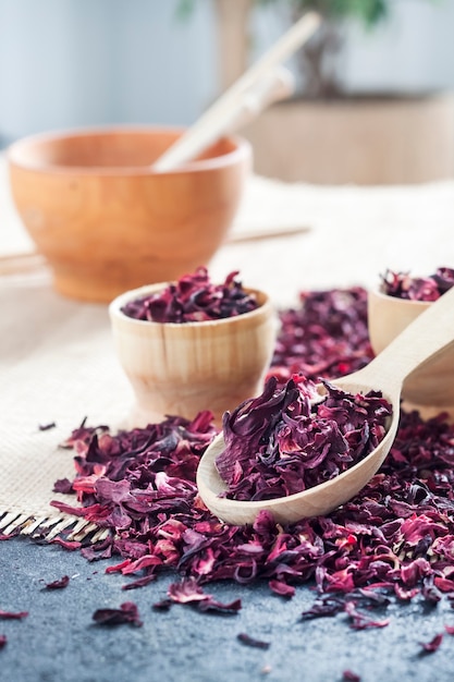 Dry hibiscus tea in wooden spoon and small cups on a table with straw mat