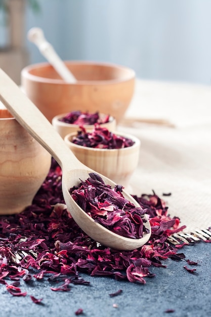 Dry hibiscus tea in wooden spoon and small cups on a table with straw mat