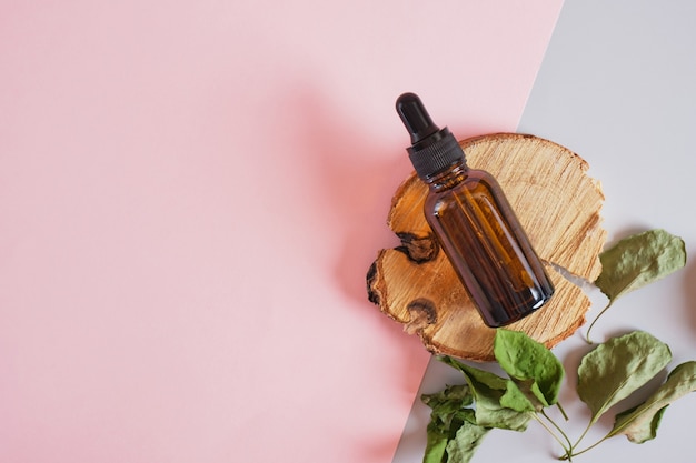 Dry leaves and brown glass bottle dropper on a podium made of wood slab on a pink background, body and face skin care, serum or cosmetic oil in a bottle with a pipette