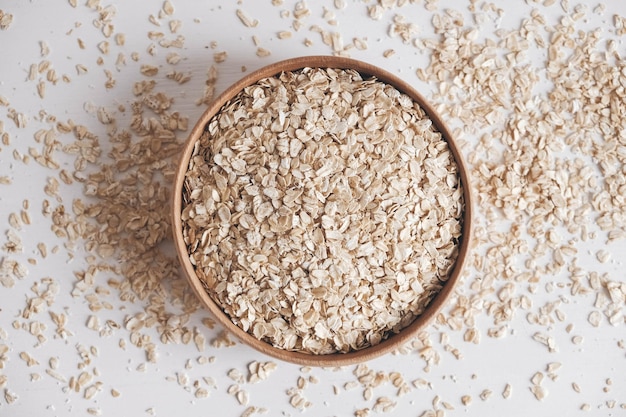 Dry oatmeal in a wooden bowl and is randomly scattered on a white background