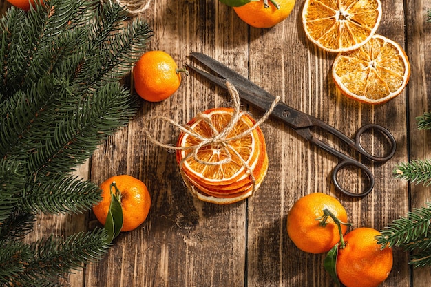 Dry oranges slices snack. Dried citrus fruits for Christmas decorations. Tangerines with leaves, fir tree branches, scissors, and thread. Wooden background, rustic concept, top view