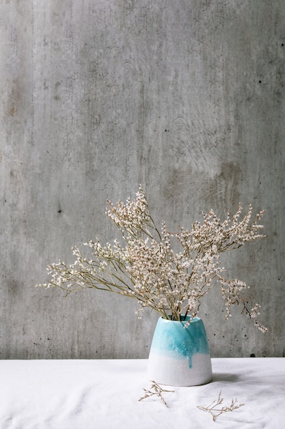 Dry white flowers branch in white ceramic vase on white linen tablecloth with gray wall behind