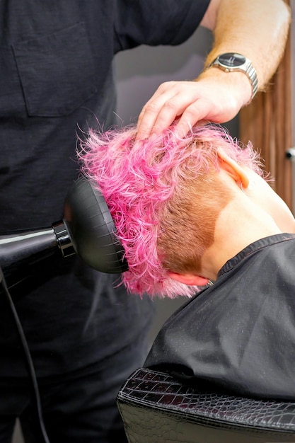 Drying short pink bob hairstyle of a young caucasian woman with a black hair dryer with the brush by hands of a male hairdresser in a hair salon close up