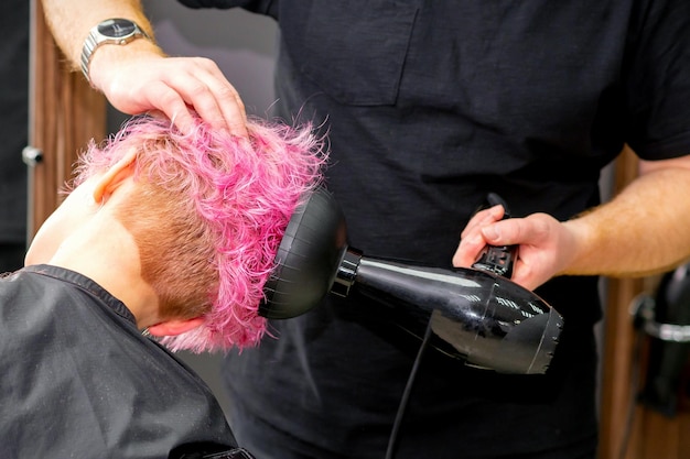 Drying short pink bob hairstyle of a young caucasian woman with a black hair dryer with the brush by hands of a male hairdresser in a hair salon close up