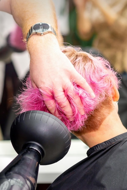 Drying short pink bob hairstyle of a young caucasian woman with a black hair dryer with the brush by hands of a male hairdresser in a hair salon close up