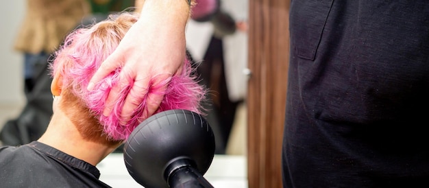 Drying short pink bob hairstyle of a young caucasian woman with a black hair dryer with the brush by hands of a male hairdresser in a hair salon close up