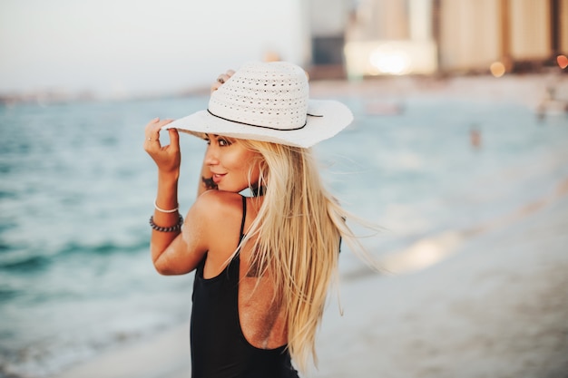 Dubai beach tourist photography. Russian lady in black bikini enjoying sunlight wearing hat best cover for Arab tourist lifestyle magazine. Overlooking blue ocean water.