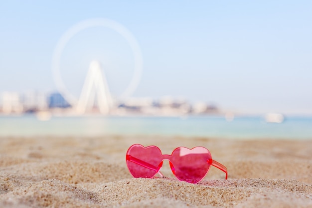 Dubai, UAE, November 2019. Pink sunglasses on a sandy beach with views of the Bluewaters Island and the Dubai Eye Ferris Wheel, honeymoon, relaxation, copy space