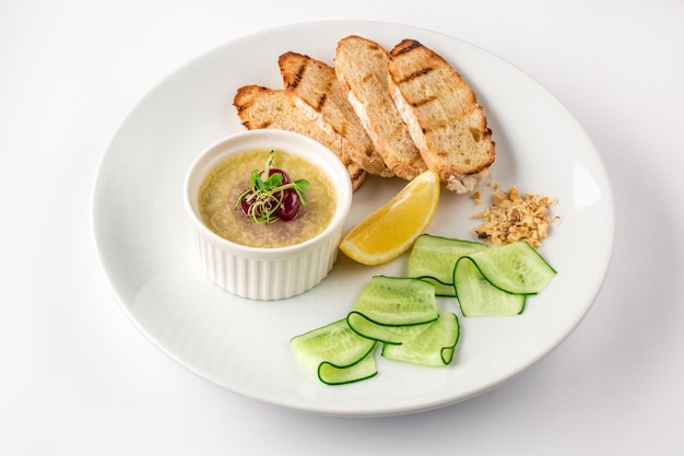 Duck liver pate with croutons, slices of lemon and thinly sliced cucumber on a white plate. Isolated on white background