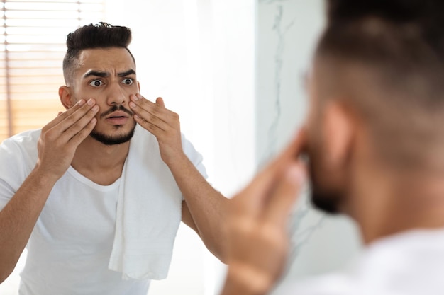 Dull skin concept worried arab guy looking at mirror and touching face