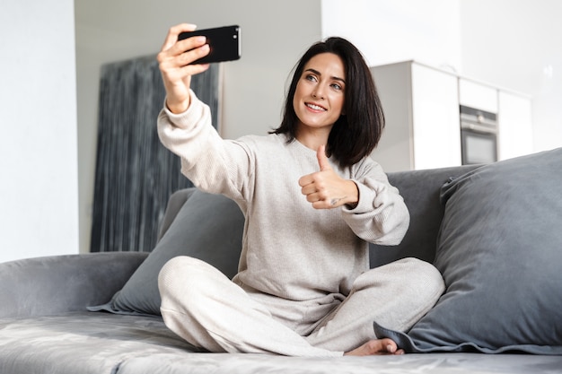 dult woman 30s taking selfie on cell phone, while sitting on couch in bright apartment