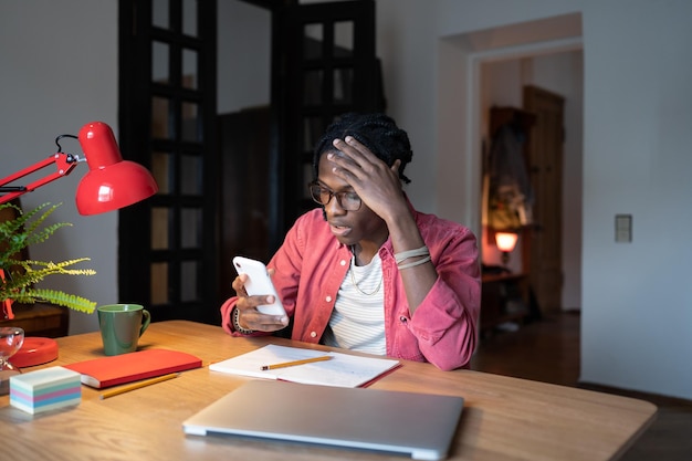 Dumbfounded confused African American man reads news in mobile phone sits at office desk with laptop