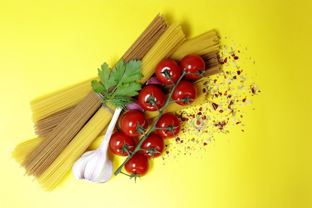 Durum wheat spaghetti and ripe cherry tomatoes, garlic and spices. Fresh greens. Bright yellow background. Products for cooking delicious pasta.