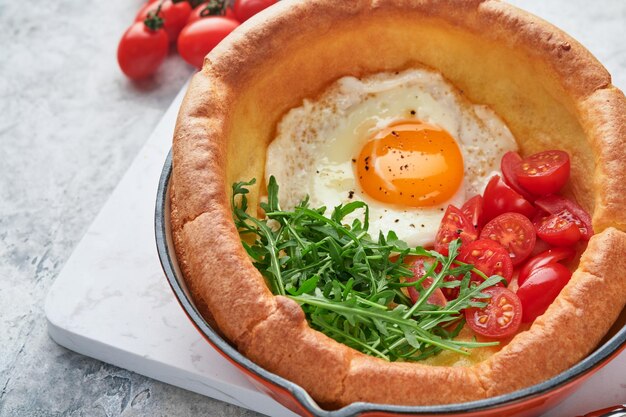 Dutch baby pancake Fresh homemade Dutch Baby pancake with fried egg tomato and green arugula in reed castiron pan on light gray concrete rustic table Flat lay close up