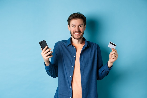 E-commerce concept. Excited young man holding smartphone and plastic credit card, shopping online, standing on blue background.