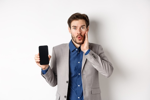 E-commerce and online shopping concept. Excited man gasping amaze and showing empty smartphone screen, wearing business suit, white background.