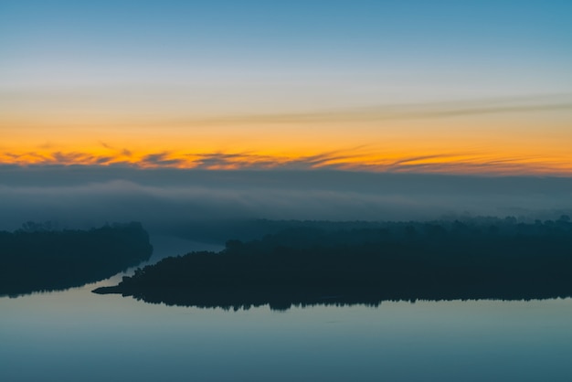 Early blue sky reflected in river water. Riverbank with forest under predawn sky. Yellow stripe in picturesque sky. Fog hid trees on island. Mystical morning atmospheric landscape of majestic nature.