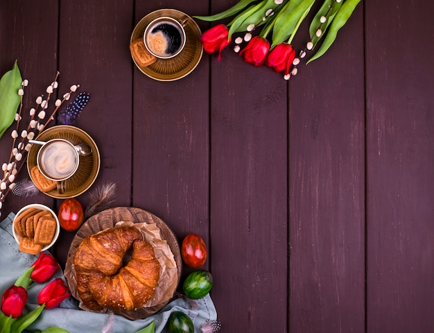 Easter breakfast with aromatic fresh espresso, croissants, colored eggs, red tulips and willow. Coffee with pastries, flowers on a wooden table. View from above. Copy space