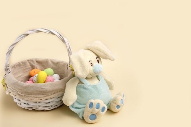 Easter bunny sits by a basket with colorful eggs on a beige background with copy space