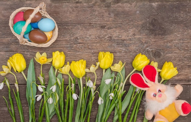 Easter card colorful eggs with a bouquet of yellow tulips and a rabbit on a wooden background with copy space