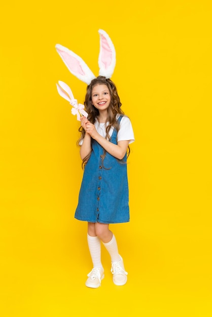 Easter for children Happy baby with long rabbit ears The spring holiday is Easter A beautiful little girl with long hair in a blue sundress fulllength on a yellow isolated background