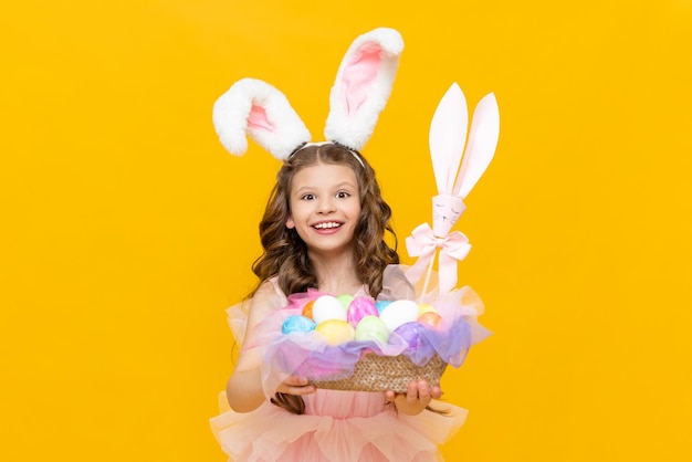 Easter for children A little girl holds a basket with colorful eggs for games on an Easter spring day Charming teenage girl with long rabbit ears on a yellow isolated background