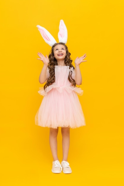 Easter children's holiday A teenage girl in a lush dress rejoices in spring The child is wearing long rabbit ears A beautiful little girl with long hair fulllength on a yellow isolated background