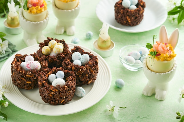 Easter chocolate nest cake with mini chocolate candy eggs with blossoming cherry or apple flowers on green background table Creative recipe for Easter table with holiday decorations Top view