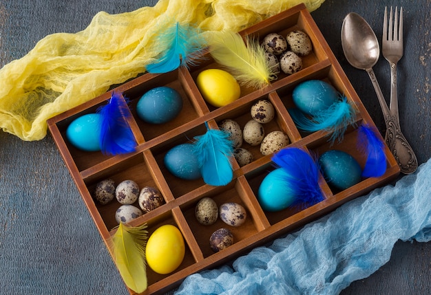 Easter composition: in the box lay blue eggs, yellow eggs, quail eggs and feathers, and cutlery - fork and spoon