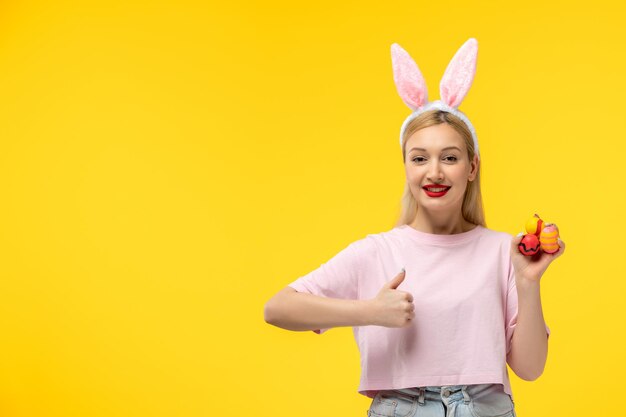 Easter cute young blonde girl wearing pink bunny ears showing good gesture and holding easter eggs