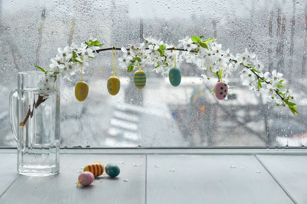 Easter eggs hanging from twig with white blossoms by the window on a rainy day