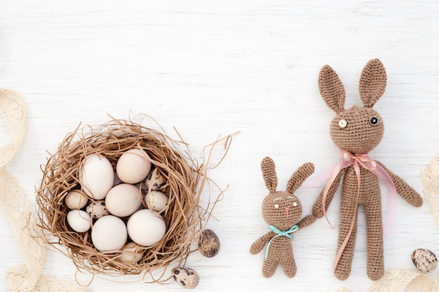 Easter eggs in nest and handmade crocheted rabbits on white background. Top view.