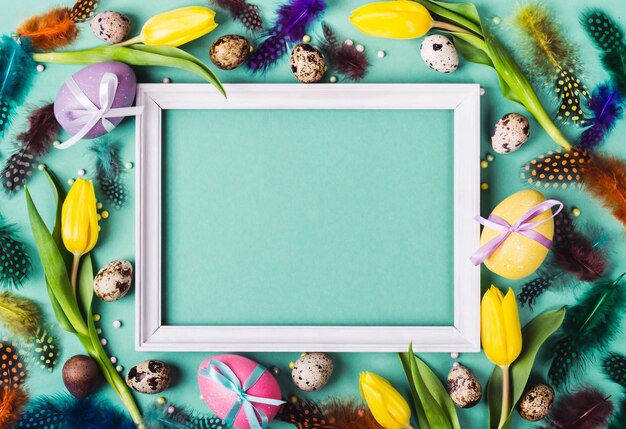 Easter Frame of yellow tulips decorated eggs and feathers on turquoise background for greeting message