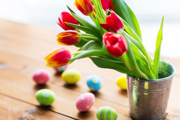 easter, holidays, tradition and object concept - close up of colored easter eggs and tulip flowers in bucket on wooden table