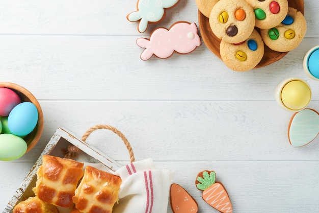 Easter hot cross buns with colored eggs Easter gingerbread rabbits and cookies over white plank wooden old table background Spring holiday baking concept Easter table setting Top view Mock up