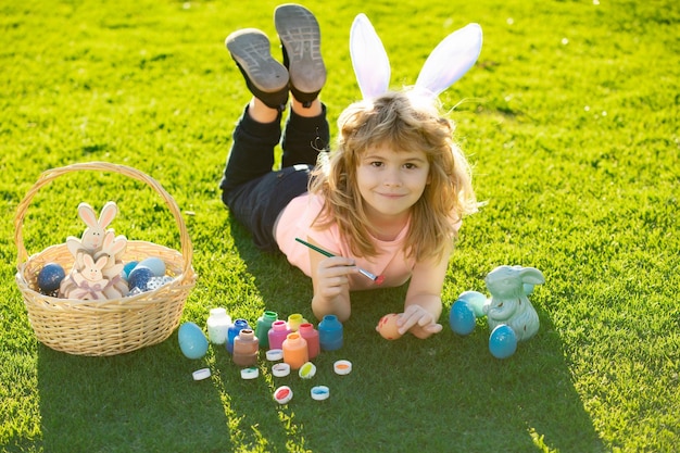 Easter kids boy in bunny ears painting easter eggs outdoor Cute child in rabbit costume with bunny ears having fun in park