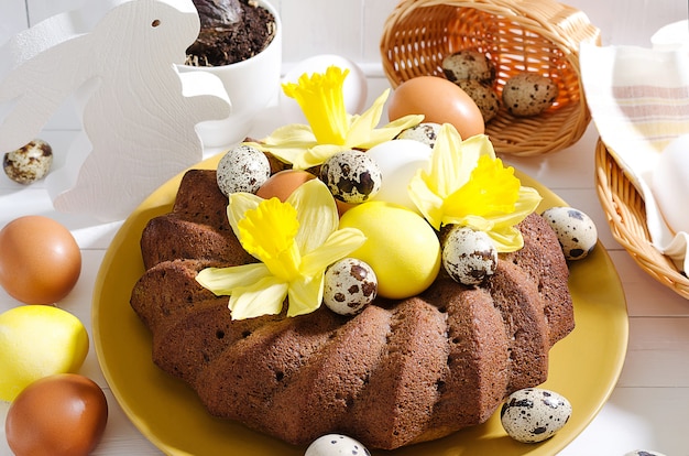 Easter still life, Easter cake with dyed eggs in a nest, daffodils on a white rustic wooden background