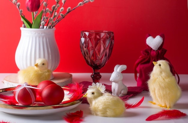 Easter still life with a white rabbit and chickens on a red background A wine glass and painted eggs the concept of the bright Easter holiday