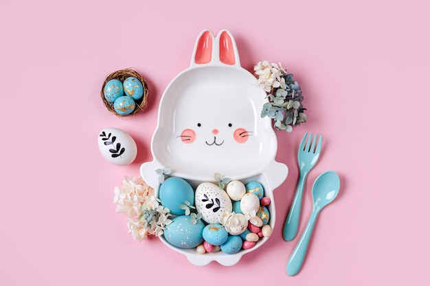 Easter table setting decoration with cute plate in the shape of a bunny with Easter eggs and candy on pink background Happy Easter concept Idea for Easter dinner